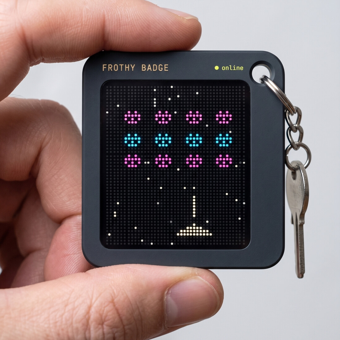 A Frothy badge — a keychain-sized dev board — held between two fingers, its dot-matrix screen lit up